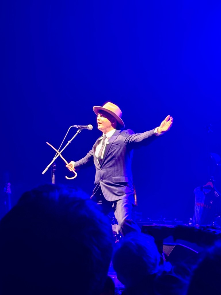 Un artiste sur scène, vêtu d'un costume à rayures et d'un chapeau, performe avec passion micro en main sous des lumières bleues.