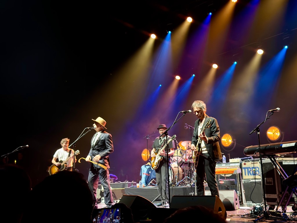 Un groupe de musiciens sur scène, jouant devant un public, avec des lumières colorées éclairant la scène.