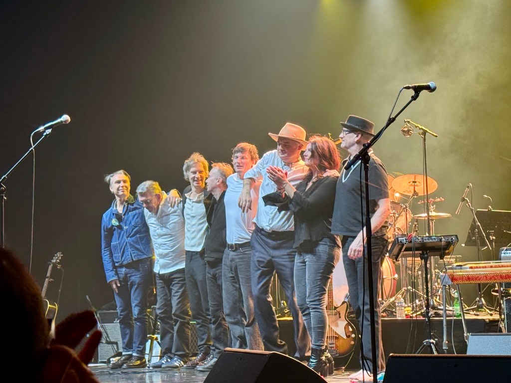 Quatre musiciens sur scène, souriants et applaudissant après une performance, avec des instruments visibles en arrière-plan.